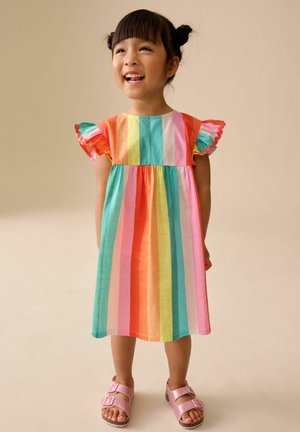 Smiling young girl with dark hair in two buns wearing a colorful striped dress and pink sandals, standing against a beige background.