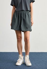 Šedé šortky Nike s elastickým pasem, s bočními rozparky a černým logem na levém stehně, doplněné bílými vysokými teniskami.