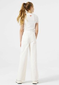 Camiseta blanca de manga corta con textura de canalé y logotipo rojo en la parte posterior, combinada con jeans blancos de pierna ancha y cintura alta. Tejido suave y corte relajado.