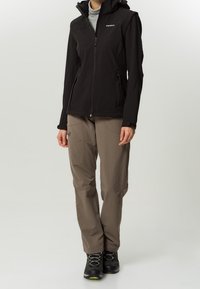 Giacca softshell nera con cappuccio, tasche con zip, abbinata a pantaloni da trekking marrone chiaro e scarponi da trekking neri con dettagli verdi.