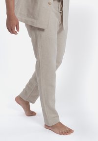 Pantaloni in lino beige chiaro, taglio dritto, con vita regolabile e tasche laterali. I piedi scalzi sono visibili mentre camminano su uno sfondo semplice.