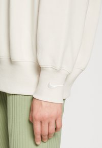 Light beige sweatshirt with ribbed cuffs and a white Nike logo on the sleeve, paired with textured green ribbed pants.