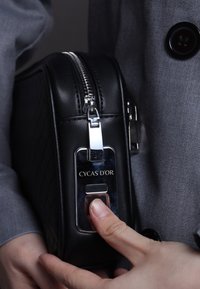 Black leather bag with diamond pattern, silver zipper, and hardware. Features a brand logo and button. Hand positioned on the button.