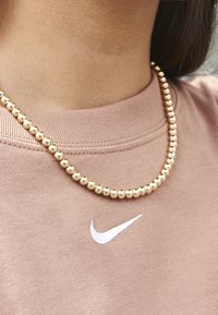 Złota naszyjnik z koralików noszony na jasnobrązowej koszuli z białym logo Nike. Koszula ma okrągły dekolt o prążkowanej fakturze.