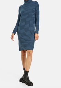 Robe à col roulé en maille bleu sarcelle, présentant un motif texturé à carreaux. Associée à des bottines noires à semelle épaisse.