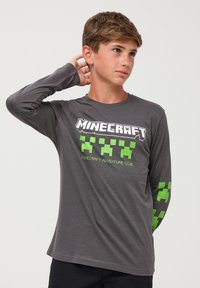 Szara koszulka z długim rękawem z nadrukiem "Minecraft Adventure Club" w białej i zielonej pikselowej grafice na klatce piersiowej i rękawach. Miękka tkanina.