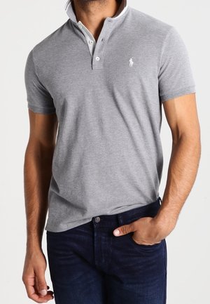 Homme portant un polo gris cintré à manches courtes avec un col blanc et un jean bleu foncé, main dans la poche.