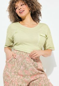 Top corto a maniche corte verde chiaro con tasca, abbinato a pantaloni larghi e stampati con disegni di animali rosa su uno sfondo beige.