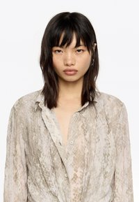 Camisa ligera y con estampado en beige con un sutil diseño de serpiente. Cuenta con un cuello, cierre de botones y un corte relajado con una tela ligeramente texturizada.