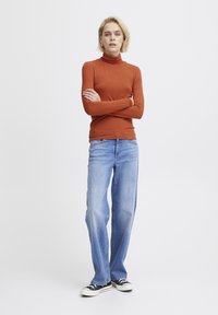 Langärmliges, rostfarbenes Rollkragenoberteil kombiniert mit ausgebleichten blauen Schlagjeans und schwarzen Low-Top-Sneakern. Glattes, strukturiertes Material.