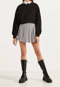 Donna che indossa un maglione corto nero sopra una camicia con colletto, una gonna plissettata a quadri e stivali neri alti fino al ginocchio con lacci, in piedi su un pavimento bianco.