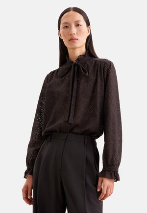 Frau trägt eine schwarze Spitzenbluse mit Schleifenkragen und Rüschen an den Manschetten, kombiniert mit hoch taillierten schwarzen Hosen.