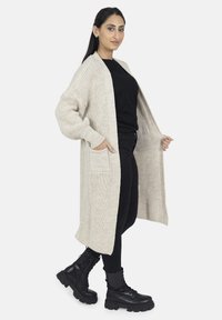 Cárdigan largo beige con un patrón de tejido texturizado, con abertura frontal, dos bolsillos delanteros y mangas caídas, combinado con pantalones negros y botas de combate.