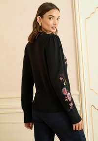 Melna adīta džemperis ar pūkainiem piedurknēm un krāsainiem ziedu izšuvumiem uz kreisās rokas, ar ribotu apakšmalu un piedurknēm.