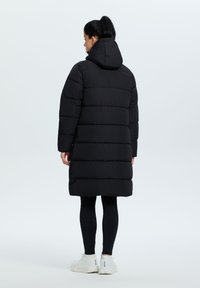Sort pufferjakke med hætte, der har vandrette quiltede sektioner, knælængde design og diskret syninger. Båret over sorte leggings og hvide sneakers.