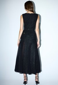 Schwarzes Abendkleid mit taillierter Taille, strukturiertem Oberteil und fließendem Rock mit transparenten Einsätzen. Akzentuiert mit subtilen Glitzer und Knöpfen.