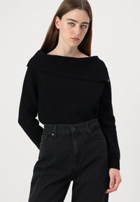 Juodas off-the-shoulder megztinis su raukšlėta tekstūra, derinamas su aukšto liemens tamsiomis džinsomis. Yra šoninė sagos detalė, suteikianti papildomo žavesio.
