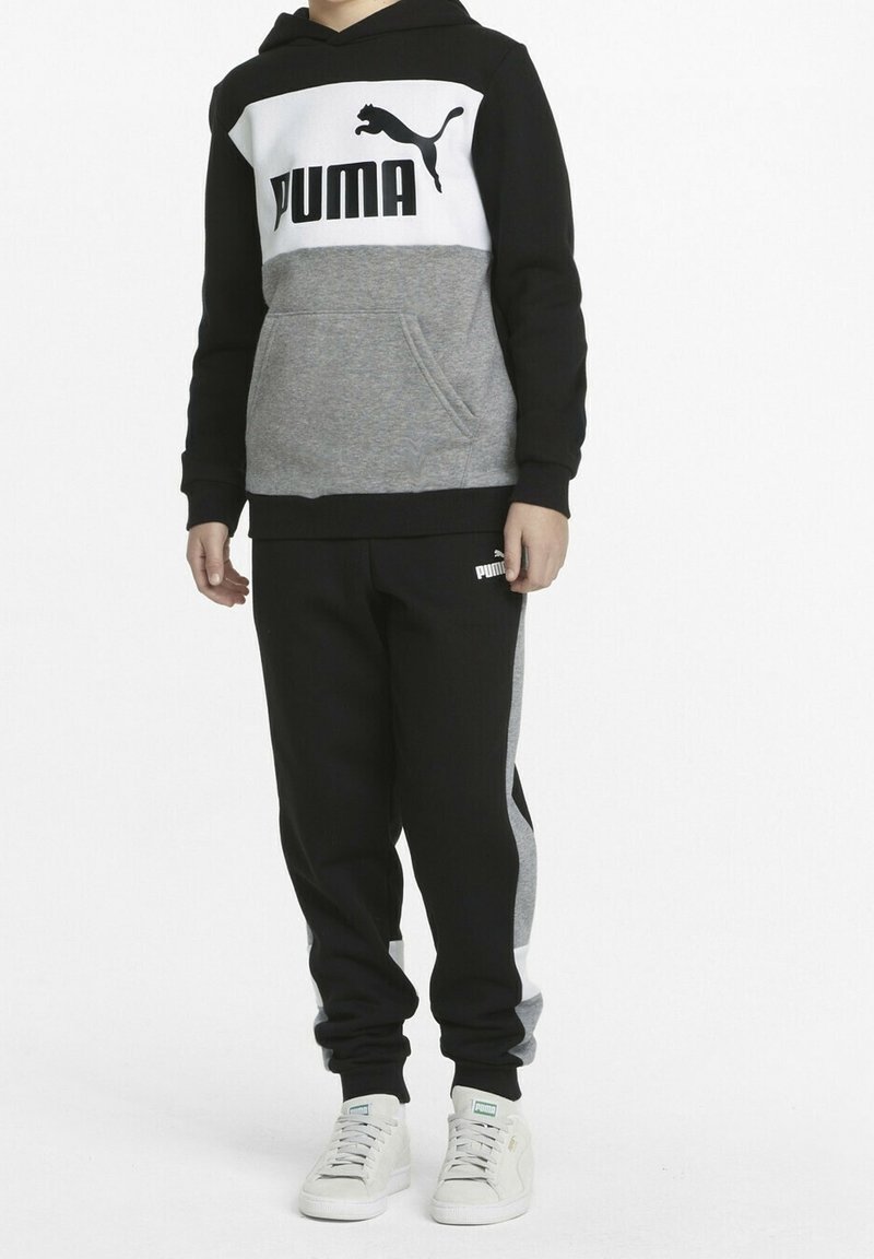 Enfant portant un sweat à capuche Puma noir, gris et blanc et un pantalon de survêtement assorti, avec des baskets Puma blanches, debout devant un fond blanc.