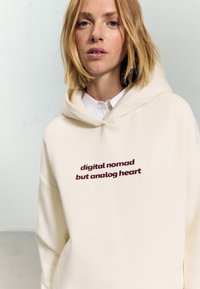 Kremowa bluza z kapturem, wykonana z miękkiego materiału, z bordowym napisem "cyfrowy nomada, ale analogowe serce", z regulowanym kapturem.