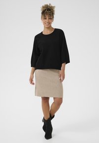 Črna pletena jopica z širokimi rokavi, kombinirana s svetlo bež svinčnik krilom. Model nosi črne gležnarje. Enostavna, čista zasnova.