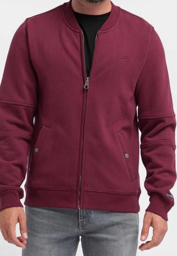 Homme portant une veste zippée bordeaux avec des poches à boutons-pression et des poignets côtelés, associée à un jean gris clair.