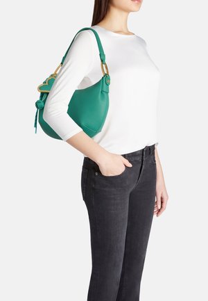 Donna che indossa una maglietta bianca a maniche lunghe e jeans neri porta una borsa a tracolla verde con accessori dorati e ciondolo a forma di cuore.