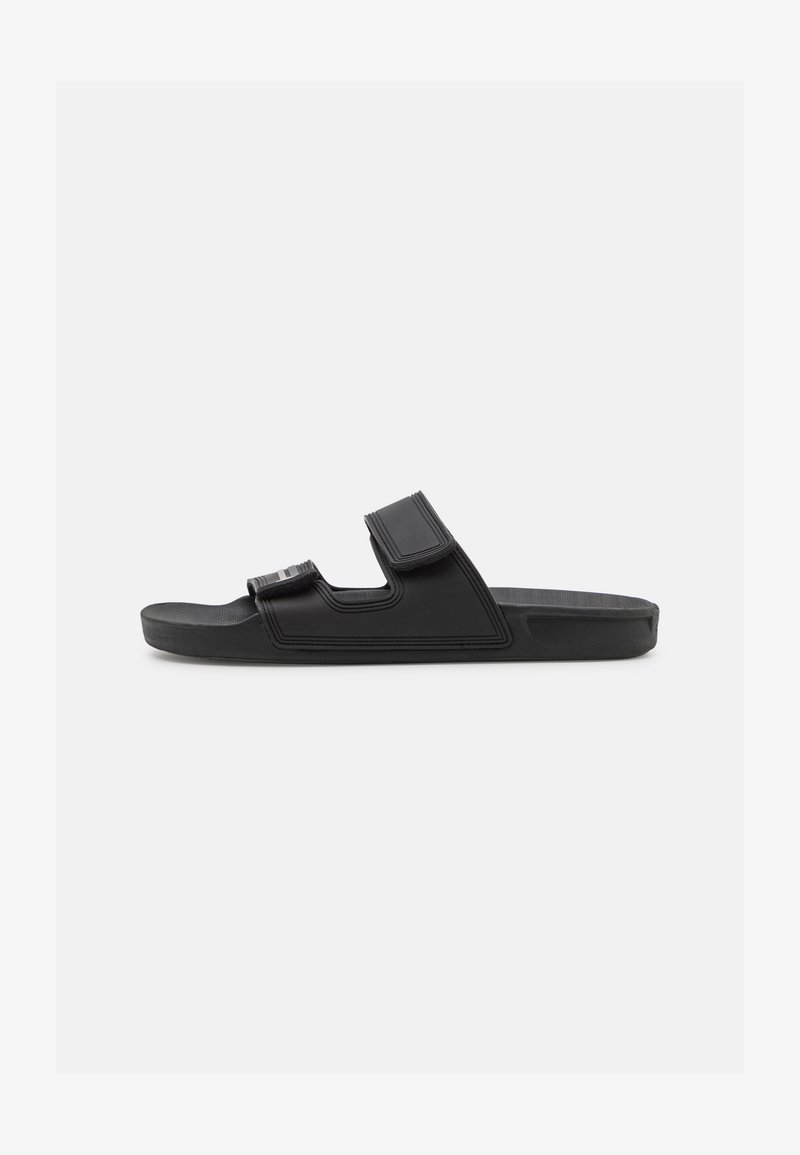 Black sandals with a flat sole, featuring a wide strap and adjustable Velcro closure. Made of synthetic material with a textured surface.