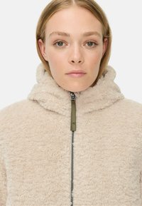 Beige, strukturiertes Fleece-Jacke mit hohem Kragen, Frontreißverschluss und kontrastierendem grünen Reißverschluss-Zieher. Weiches, gemütliches Material.