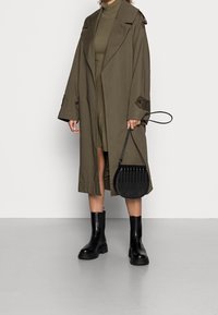 Trench coat verde oliva con vestibilità oversize e tasche laterali, abito a coste verde oliva, stivaletti neri alla caviglia e borsa rotonda in tessuto nero.