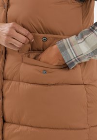 Bruine puffer vest met een gestikte textuur, voorzien van drukknopen en zijzakken. Een mouw van een geruite shirt is zichtbaar in de afbeelding.