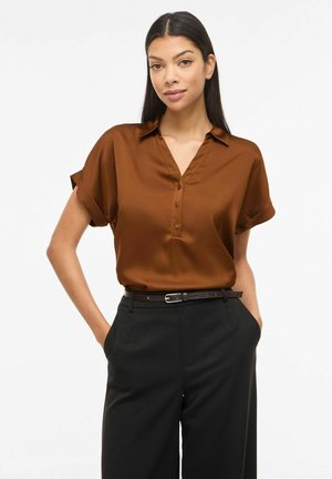 Femeie cu păr lung purtând o cămașă maro cu mânecă scurtă cu nasturi, băgată în pantaloni negri cu o curea neagră, stând cu mâinile în buzunare.