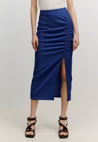 Mittelblaue, strukturierte Midirock mit Raffung an einer Seite und einem hohen Schlitz bis zum Oberschenkel. Kombiniert mit braunen, geschnallten, hohen Sandalen.