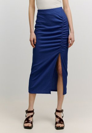 Blauwe midi-rok met textuur, voorzien van geplooide details langs één kant en een hoge split tot de bovenbeen. Gecombineerd met bruine strappy haksandalen.