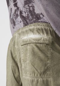 Lichte olijfgroene pantalons van zachte stof, met een achterzak en een elastische tailleband voor comfort. Geverfde afwerking met subtiele glans.