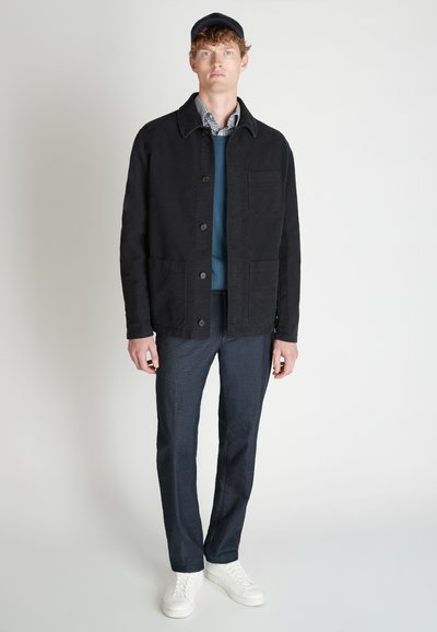 Chaqueta de algodón negra con cierre de botones y dos bolsillos delanteros, superpuesta sobre un suéter azul. Combinada con pantalones oscuros y zapatillas blancas.
