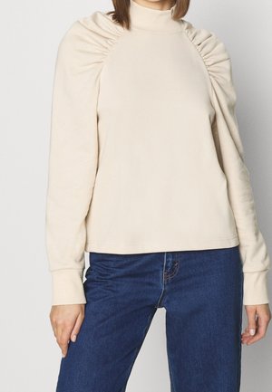 Pullover beige con collo alto, caratterizzato da dettagli di tessuto arricciato sulle spalle e polsini aderenti, abbinato a jeans blu scuro.