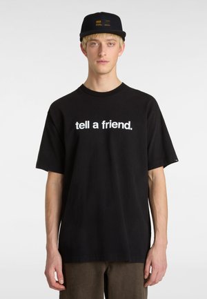 Black cotton t-shirt featuring a white graphic print reading "tell a friend." Model is wearing a black cap with yellow accents.