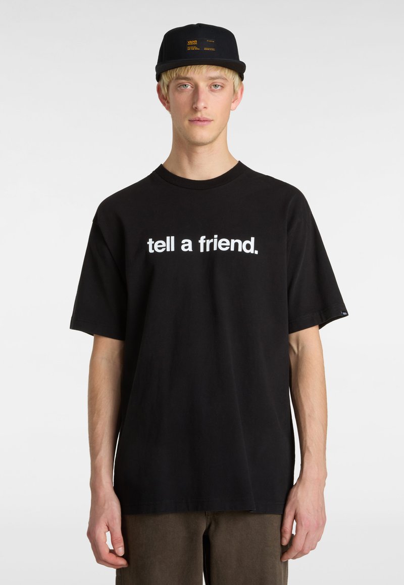 Sort bomulds t-shirt med et hvidt grafikprint, der står "fortæl en ven". Modellen bærer en sort kasket med gule accenter.