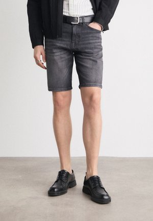 Musta suorat leikatut denimshortsit, yhdistettynä mustiin nauhallisiin kenkiin. Materiaalissa on kevyt kiilto, näkyvissä ompelujen yksityiskohdat taskuissa.