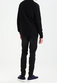 Person som står med ryggen vänd, iförd svart långärmad tröja, svarta jeans och mörkblå sneakers med vita sulor mot en neutral bakgrund.