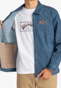 Denim-takk med lys blå vask, med et brodert logo på brystet, og et mønstrete innvendig fôr. Hvit t-skjorte under.