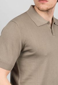 Beige polo tričko s krátkým rukávem vyrobené z ribbovaného pleteného materiálu. Obsahuje klasický límec a zapínání na dvě knoflíky, s přiléhavým střihem.
