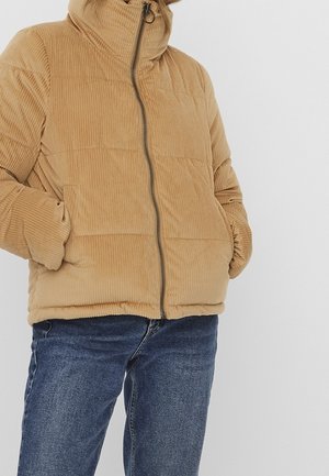 Personne portant une doudoune en velours côtelé beige avec fermeture éclair à l'avant et mains dans les poches, associée à un jean en denim bleu.