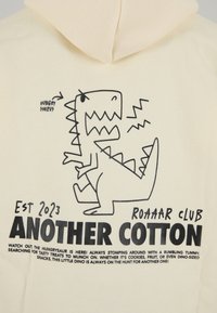 Sudadera con capucha color crema con un gráfico negro de un dinosaurio con dientes exagerados y el texto "HUNGRYSAURUS" y "ANOTHER COTTON" en negrita.