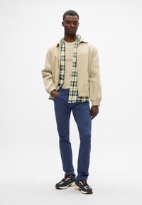 Beżowa kurtka bomber, koszula w kratę nałożona na beżową bluzkę, niebieskie spodnie i czarno-beżowe trampki z akcentami. Model stoi na tle białym.