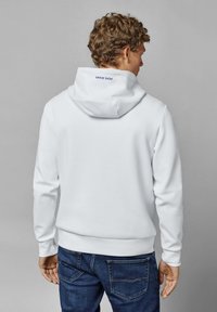 Sudadera blanca con bolsillo canguro, puños y dobladillo acanalados. Presenta un logo azul en la parte posterior de la capucha. Llevada con jeans azules.