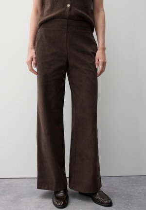 Persoon die donkerbruine corduroy broek met hoge taille en wijde pijpen draagt, met zwarte loafers, staand tegen een effen muur.