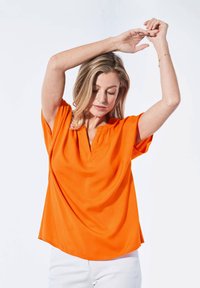 Kurzärmliges, lockeres oranges Oberteil mit V-Ausschnitt und weichem Stoff. Getragen mit hellen Hosen vor einem einfarbigen Hintergrund.