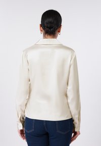 Μπλούζα από σατέν σε χρώμα μαργαριταριού με κλασικό γιακά, μακριά μανίκια και μανσέτες με κουμπιά, χαρακτηρισμένη από λεία, γυαλιστερή υφή.