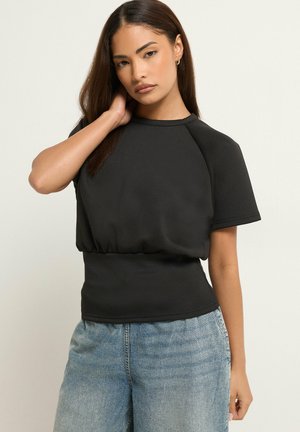 Kvinde med langt brunt hår iført en sort kortærmet top og lyseblå jeans, poserer med den ene hånd, der rører ved hendes nakke